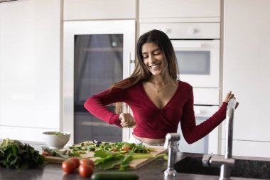 Neşeli, rahat, genç, yetişkin Hintli kız ev mutfağında müzik yapıyor, organik gıda malzemeleriyle masada dans ediyor, yeşil sebzeleri kesiyor, eğleniyor, sağlıklı yaşam tarzının tadını çıkarıyor.