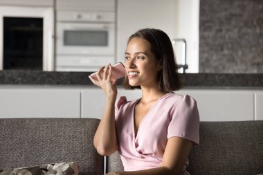 Pozitif genç cep telefonu kullanıcısı kız, elinde aletle, sesli mesaj dinliyor, ses iletişiminin tadını çıkarıyor, akıllı telefonuyla kanepede oturuyor, başka tarafa bakıyor, gülümsüyor.