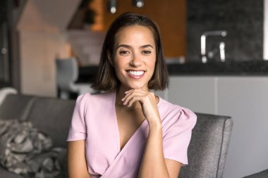 Mutlu, genç, güzel, 20 'li yaşlarda beyaz bir kadın evde portre için poz veriyor, oturuyor, koltukta dinleniyor, çenesine dokunuyor, mükemmel dişlek gülüşüyle kameraya bakıyor. Pozitif kadın model portresi