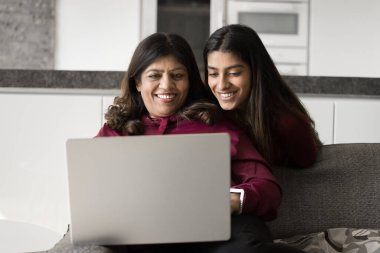 Mutlu Hintli bir anne ve neşeli bir kız çocuğu uygulamayı kullanıyor, bilgisayarla internet üzerinden iletişim kuruyor, bilgisayarları birlikte kullanıyor, evde kanepede oturuyor, aletlerle boş vakitlerin tadını çıkarıyor.