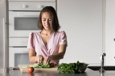 Salata için taze sebze doğrayan, tahtanın üzerine yeşil yapraklar kesen, organik malzemelerden sağlıklı yemekler yapan çekici genç bir ev hanımı.