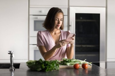 Odaklanmış genç aşçı blogcu kadın cep telefonuyla yazı yazıyor, taze sebzelerin üzerine cihaz koyuyor, pnline marketten organik gıda malzemeleri sipariş ediyor.