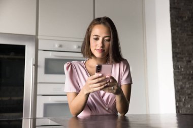 Cep telefonunda akıllı ev uygulaması kullanan, arka planda aletlerle mutfak masasında duran, daktilo kullanan, internet iletişiminden keyif alan genç, güzel bir kız.