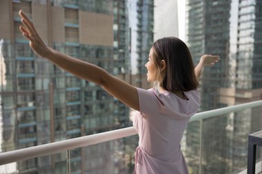 Neşeli genç ev sahibi kadın şehir merkezinin balkonunda duruyor, şehir merkezine bakıyor, açık kollarla, temiz hava soluyor, neşe, mutluluk, büyük şehirde yaşamaktan keyif alıyor.