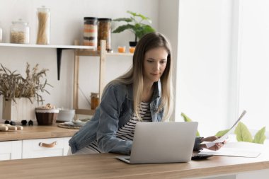 Genç, odaklanmış bir kadın mutfakta duruyor, muhasebe işi yapıyor, kağıt döküman tutuyor, ev faturalarını kontrol ediyor, modern banka başvurularını laptopta kullanıyor, evrak işleriyle meşgul, bilgi karşılaştırıyor.