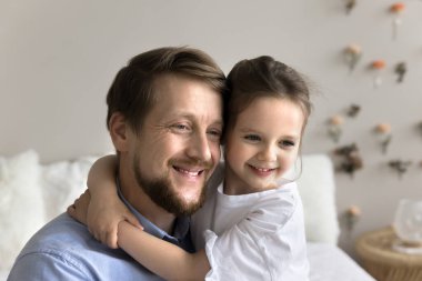 Sevimli anaokulu öğrencisi 4 'ün kızı sarılıyor genç baba gülümsüyor başka tarafa bakıyor, yakın çekim. Aile birlikte vakit geçirir, kucaklaşır, iletişimden zevk alır, sıcak ilişkileri olur, koşulsuz sevgiyi hissederler.