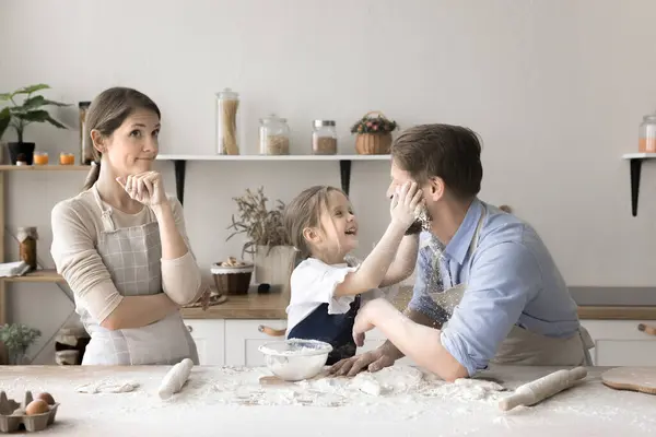 stock image Young woman in apron feels bewilderment while looks at mess in kitchen while her husband and little daughter cooking homemade pastries in chaos, fooling, have fun stained with white flour. Family fun