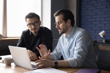 İki genç odaklı iş ortağı modern ofiste masa başında oturmuş proje detaylarını tartışıyor, fikirleri paylaşıyor, çalışanlara yeni iş uygulamaları öğretiyor ve online çalışmaları açıklıyor. İşbirliği, takım çalışması