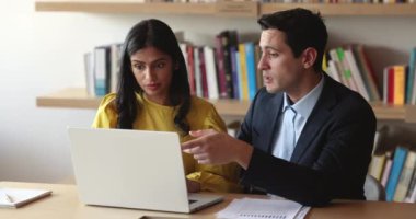 Birlikte bilgisayar üzerinde çalışan çok ırklı iş arkadaşları, internet projesini, çevrimiçi içeriği, takım çalışmasını, ofiste dizüstü bilgisayarda yazı yazmayı ve arka planda kütüphane kitaplıklarını tartışıyorlar.