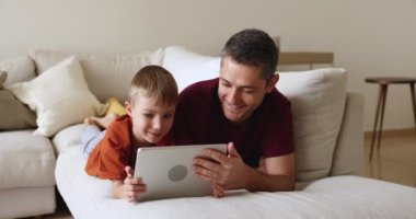 Orta yaşlı mutlu bir baba ve küçük tatlı bir çocuk internet uygulamasını tablette kullanıyor, evde kanepede dinleniyor, öğrenme içeriğini izliyor, bir aletin üzerinde sanal aile oyunu oynuyor, online iletişimden keyif alıyor.