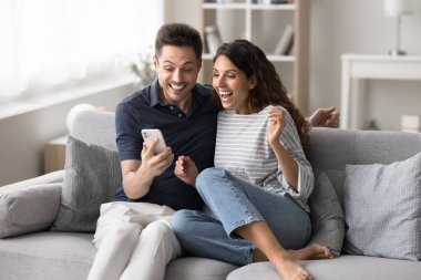 Neşeli Latin eşler akıllı telefondan harika haberler okur, koltuğa oturur, piyango zaferinin tadını çıkarır, ikramiyeyi kazanma anını kutlar, inanılmaz mutlu ve şaşkın görünürler. Banka kredisi onayı, satış, büyük indirimler.