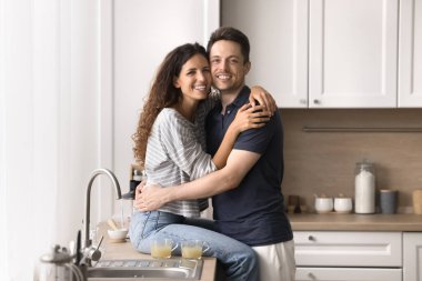 Güzel evli çift mutfakta kameraya poz veriyor. Mutlu İspanyol kadın tezgahta oturur, yakışıklı kocasına sarılır ve kameraya bakar. Ev sahipleri aile portresi, aşk, ilişkiler