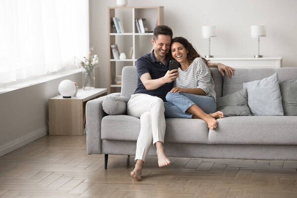 Счастливые супруги проводят беззаботные выходные дома, сидя на диване, держать мобильный телефон, смотреть смешные он-лайн видео, смеяться, наслаждаться забавным содержанием интернета, имея приятный разговор с родственником через видео-звонок