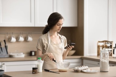 Kadın akıllı telefonu mutfak masasında tutuyor, yemek pişirme uygulaması kullanıyor, internette yemek tarifleri okuyor, yumurta çırpıyor, nefis ev yapımı krepler hazırlıyor, yemek blogcusu sosyal ağda fotoğraflarını paylaşıyor.