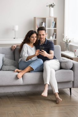 Neşeli aile çift oturma odasındaki kanepede dinleniyor, cep telefonunu eğlence için kullanıyor, internetten alışveriş yapıyor, alışverişi tartışıyor, ev için ürünler seçiyor, sosyal ağlara takılıyor, tembelce hafta sonunun tadını çıkarıyor.