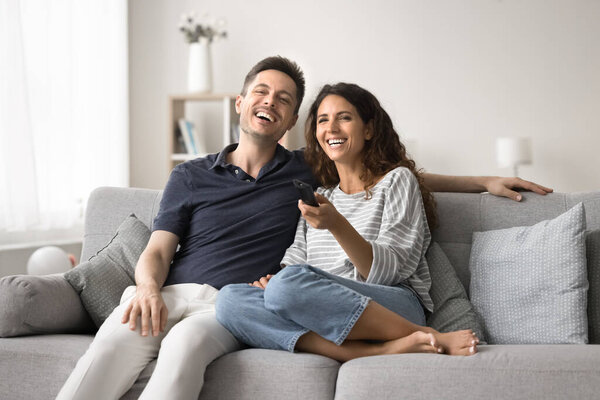 Смеющаяся латиноамериканская пара удобно сидит на диване, поглощенная просмотром телевизора, держит пульт дистанционного управления, переключает каналы, выбирает свое любимое шоу, регулирует громкость, расслабляется вместе в выходные дни дома