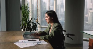 Dizüstü bilgisayarda çalışan, müşteriyle internette sohbet eden, modern ofiste tekerlekli sandalyede oturan ciddi İspanyol kadın çalışanı. Uzak meslek, teknoloji, iş akışı