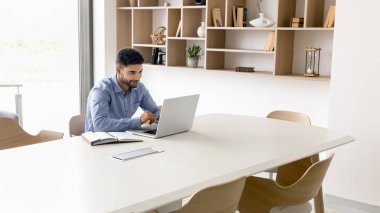 Ofisteki ortak çalışma odasında bilgisayar başında çalışan, dizüstü bilgisayarda yazan, büyük toplantı masasında tek başına oturan, gülümseyen, çevrimiçi iletişimden keyif alan genç, meşgul bir Arap proje yöneticisi.