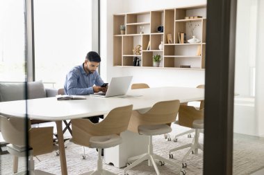 İşletme iletişimi için dijital cihaz kullanan odaklanmış genç bir Arap çalışanı, ofis çalışma alanında tek başına büyük bir masada oturuyor, dizüstü bilgisayarında pn smartphone yazıyor.
