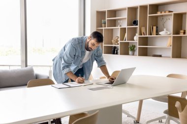 Odaklanmış girişimci adam aceleyle çalışıyor, dizüstü bilgisayarla yazı yazıyor, çalışma odasında notlar yazıyor, büyük işyeri toplantı masasında duruyor. Yetişkin öğrenci çevrimiçi bilgisayar dersini izliyor