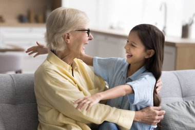 Neşeli çocuk kız ve mutlu büyükanne kucaklaşmak, kucaklamak, gülmek, evde kanepede oturmak, aile etkinliğini kutlamak için kollarını açıyorlar. Büyükanne ve torun sevgiyle sarılıyor.