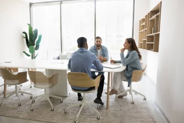 Üç kişilik küçük işletme ekibi iş görüşmesi için büyük bir masada oturuyor, proje üzerinde beyin fırtınası yapıyor, işbirliği planlıyor. Ofis beyaz iç mekan, cam duvar manzaralı.