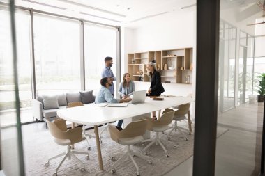 Küçük genç iş grubu ve yaşlı lider kadın modern ofis alanındaki büyük masada konuşuyor, toplantı odasının içinde, projeyi tartışıyor, gülüyor, beyin fırtınası yapıyor, takım çalışmasından keyif alıyor.