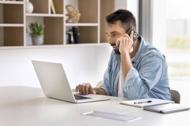 Laptop 'ta cep telefonuyla konuşan yakışıklı Latin işadamı iş iletişimi için kablosuz internet teknolojisini kullanıyor, telefonda müşterilere danışmanlık yapıyor.