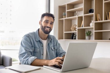 Ofis masasında dizüstü bilgisayarda yazan, dişlek gülümsemeyle kameraya bakan, profesyonel portre için poz veren, iş başarısının keyfini çıkaran, başarılı bir iş yapan yakışıklı Latin girişimci.
