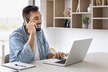 Neşeli, yakışıklı İspanyol girişimci iş yerinde cep telefonuyla konuşuyor, dizüstü bilgisayarda yazıyor, gülümsüyor, sohbete gülüyor, çoklu iş iletişiminden keyif alıyor.