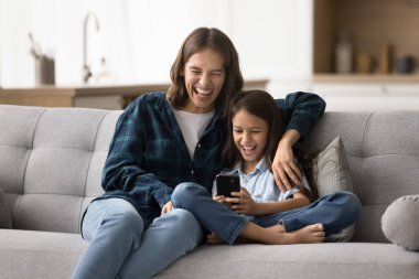 Neşeli bir anne akıllı telefondan kızın online oyun oynamasını izliyor. Mutlu anne-kız çocuğu cep telefonunu eğlence için kullanıyor, sesli gülüyor.