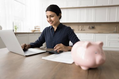 Tatmin olmuş Hintli kadın evde muhasebecilik yapıyor, mali rapor hazırlıyor, elektronik banka başvurusu ile ev faturalarını ödüyor, kar, gelir ve kazancın sembolü olarak masadaki domuz kumbarası