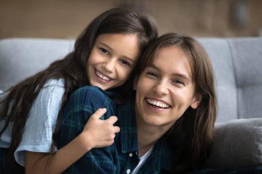 Neşeli genç anne ve pozitif kız, kanepeye yığılmış, dişlek tebessümle kameraya bakıyor, gülüyor, ev portresi için poz veriyor, aktif oyunlardan, yakınlıktan, boş zamanlardan keyif alıyor.