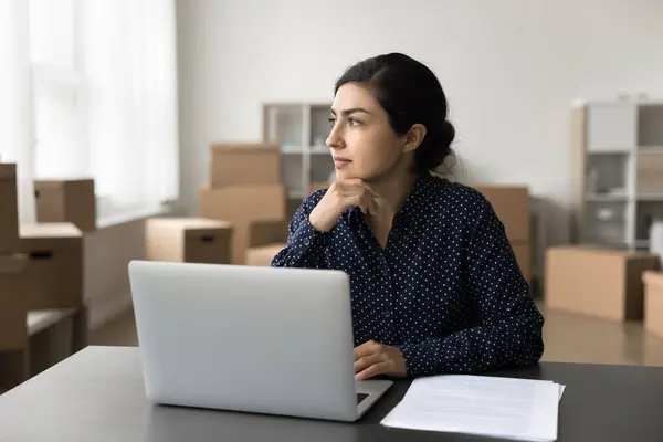 stock image Pensive Indian woman sit at desk with laptop, thinking on marketing campaign development or ideas, working in warehouse, heap of cardboard boxes on background. Drop shipping, electronic commerce, SEO