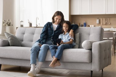 Neşeli, güzel, genç bir anne, cep telefonu kullanarak kızını kucaklıyor, cep telefonundan internet içeriğine bakıyor, gülümsüyor, gülüyor, annelikten keyif alıyor, aletle aile eğlencesi yapıyor.