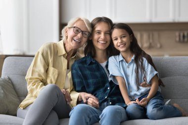 Üç nesildir aile portresinin mutlu kızları ve kadınları. Neşeli kız, anne ve büyükanne rahat koltukta oturuyor, sarılıyor, dişlek tebessümle kameraya bakıyor.