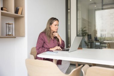 Yaratıcı proje üzerinde çalışan odaklanmış iş kadını toplantı odasında büyük bir ofis masasında tek başına oturuyor, çeneye dokunuyor, bilgisayarda düşünüyor, ekranda metni okuyor.