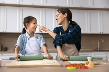 Neşeli genç anne ve sevimli yardımcı kız çocuk yemek pişiriyor ve oynuyor. Mutlu anne parmaklarıyla çocuğun yüzüne dokunuyor, burnunda süslü bir leke bırakıyor, gülüyor, kızlara pişirmeyi öğretiyor.