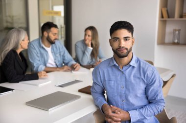 Ciddi yakışıklı genç Arap CEO 'su arka planda çalışan çeşitli iş ekibiyle toplantı masasında oturuyor, kameraya bakıyor, profesyonel portre için poz veriyor, liderliği destekliyor.