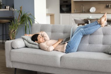 Kaygısız Hintli kadın internette tembellik edip rahat bir kanepede dizüstü bilgisayarla yatar, web sörfü bilgileri, internet sitesi, ucuz biletler, elektronik ticaret hizmetleri kullanarak otel rezervasyonları yapar.