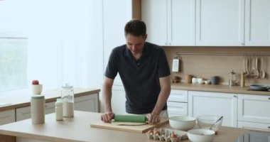 Olumlu yakışıklı genç aşçı ev mutfağında fırın yemeği hazırlıyor, hamur hamurunu hamur tahtasının üzerinde pasta malzemesiyle yuvarlıyor, şenlikli aile yemeği için fırında tatlı pişiriyor.