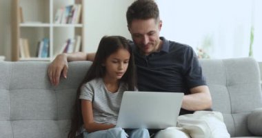 Odaklanmış bir baba ve dizüstü bilgisayarda oynayan tatlı ergen bir kız çocuğu, kanepede birlikte oturuyor, yazışıyor, neşeli oluyor, gülüyor, internetteki aile eğlencesi için alet kullanıyor.