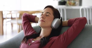 Modern kablosuz kulaklıklarla müzik dinleyen mutlu kadın ellerini başının arkasına koy, rahat bir kanepeye uzan, evde yalnız başına, kapat. Dijital akış hizmetleri abonelik hizmetleri
