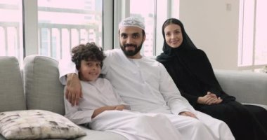 Neşeli, genç, Arap ebeveynler ve evde oturan sevimli bir çocuk aile fotoğrafı için poz veriyor. Mutlu Müslüman çift Abaya giyer, çocuğa sarılır, kameraya bakar, güler.