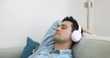 Huzurlu, uykulu, kablosuz kulaklıklı, rahat koltukta dinlenen, sakin müzik dinleyen, boş zamanların tadını çıkaran, online multimedya listesindeki favori melodileri çalan İspanyol genç adam.