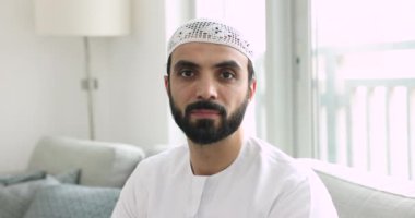 Beyaz Arap kıyafetleri giymiş yakışıklı Müslüman bir adam, kameraya bakıyor, mutlu, pozitif, gülümsüyor, evinde portre pozu veriyor, evinde koltukta oturuyor.