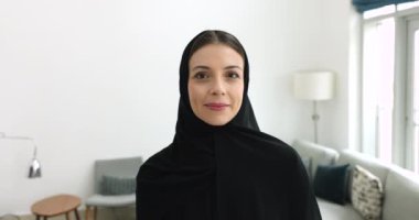Geleneksel siyah başörtüsü takan çekici Arap kadın ve abaya evde poz veriyor, kameraya bakıyor, gülümsüyor, gülüyor, mutlu oluyor, neşeli oluyor, yüzünü çeviriyor, düşünüyor.