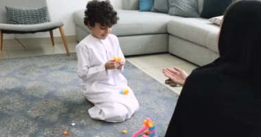 Beyaz etek giymiş tatlı Arap çocuk ve siyah abaya giyen anne ev zemininde oyuncak tuğlalarla oynuyor renkli blokları diziyor, eğleniyor, aile oyunlarından keyif alıyor, eğleniyor.