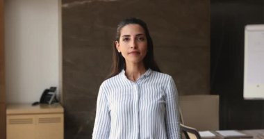 Ciddi, güzel, genç, İspanyol iş adamı bir kadın ofiste duruyor, kendine güven jesti yapıyor, mutlu oluyor, dişlek bir gülümsemeyle kameraya bakıyor. CEO, yönetici, yönetmen vesikalığı.