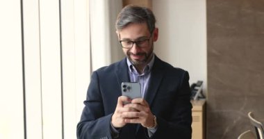 Pozitif orta yaşlı İspanyol iş adamı cep telefonuyla yazı yazıyor, cep telefonuyla ofis penceresinde duruyor, internette sohbet ediyor, internet üzerinden iş iletişimi için cihaz kullanıyor.